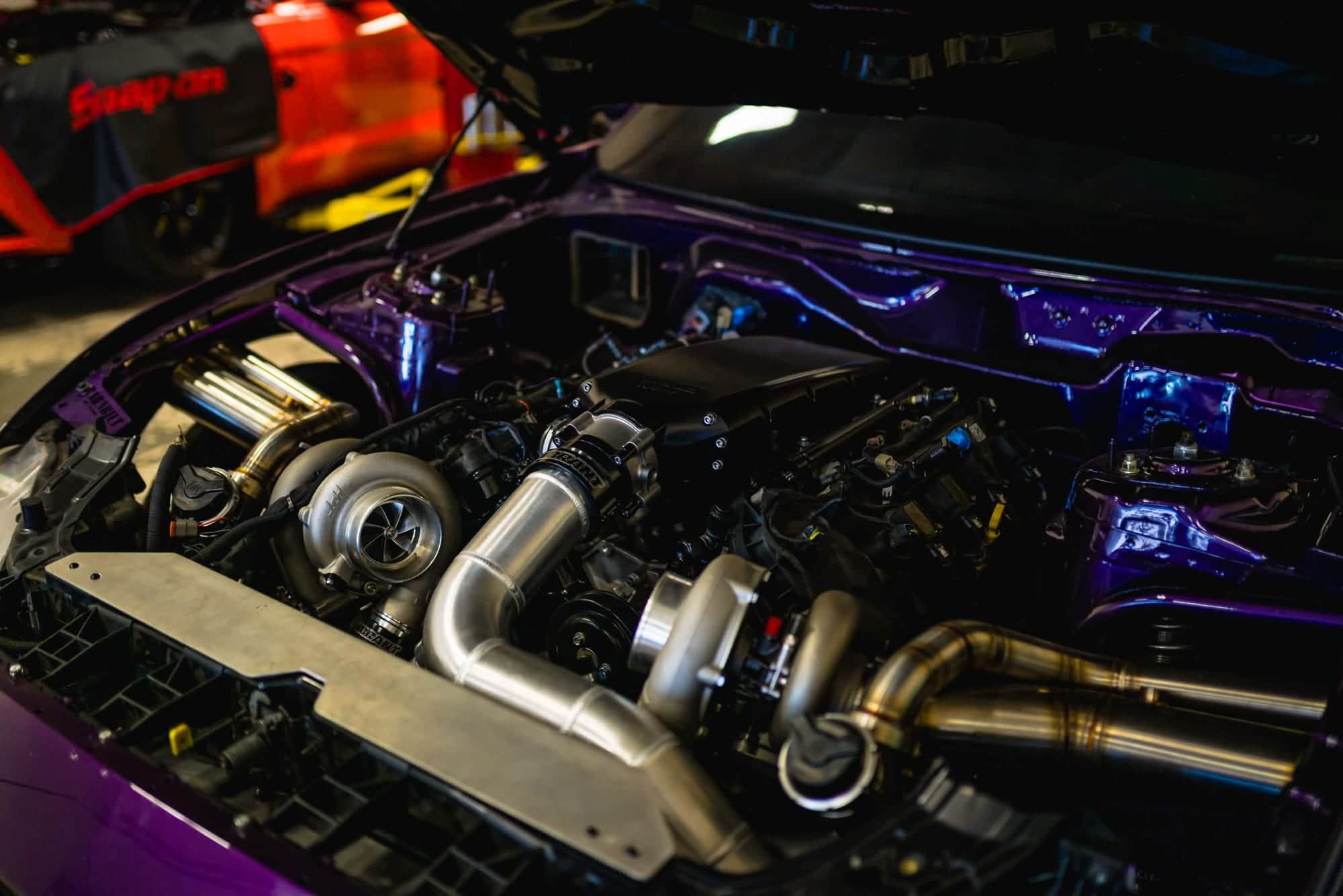 Engine bay of a purple Mustang with twin turbochargers, polished charge piping and stainless hot side