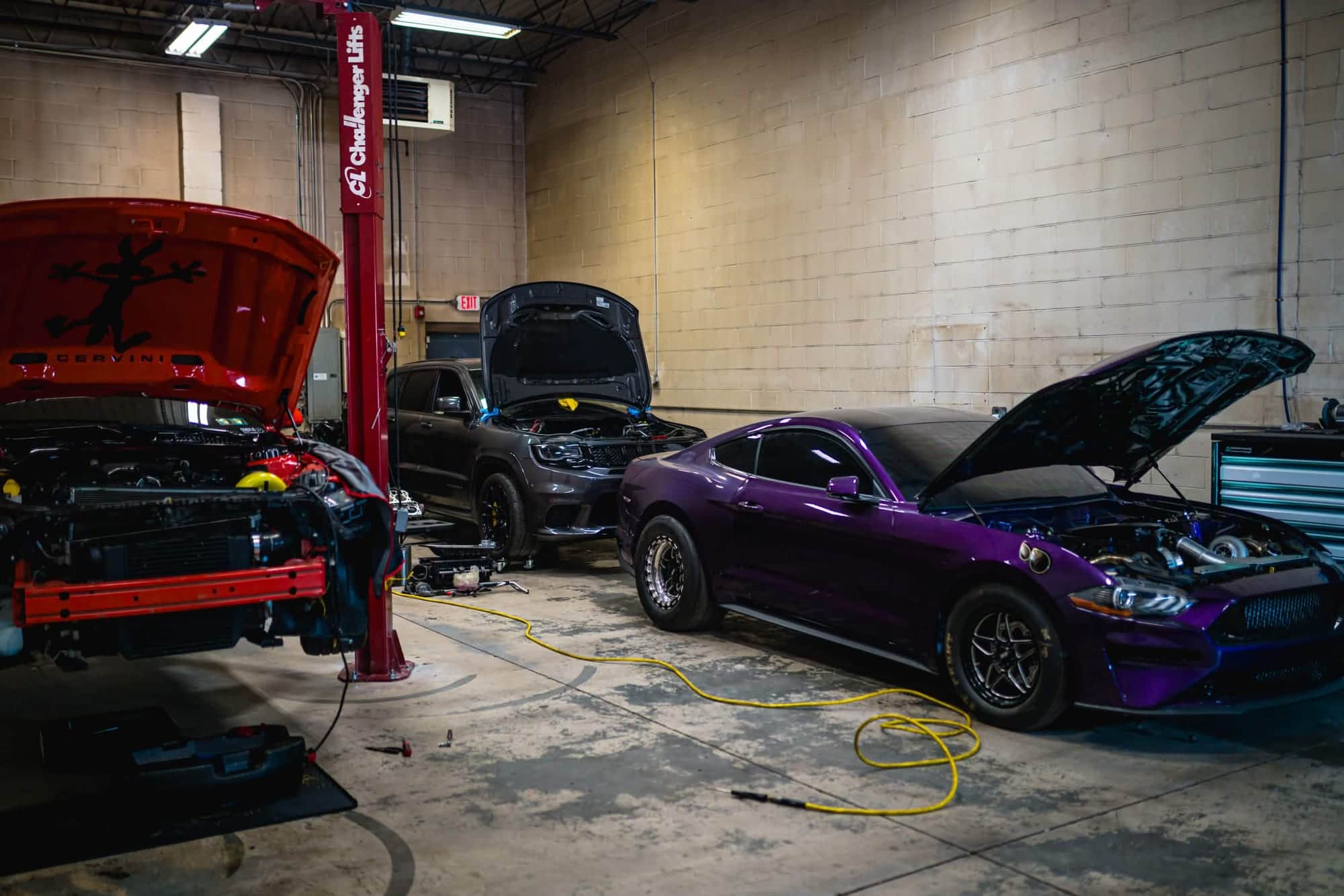 The Noro Performance shop floor with a red Mustang on the lift, a Trackhawk and a purple twin-turbo Mustang, all with hoods up