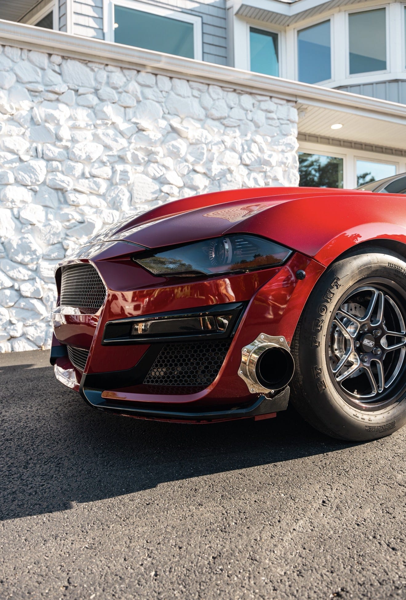 Front corner of a red Mustang showing the polished turbo outlet exiting through the front bumper and a skinny front drag wheel