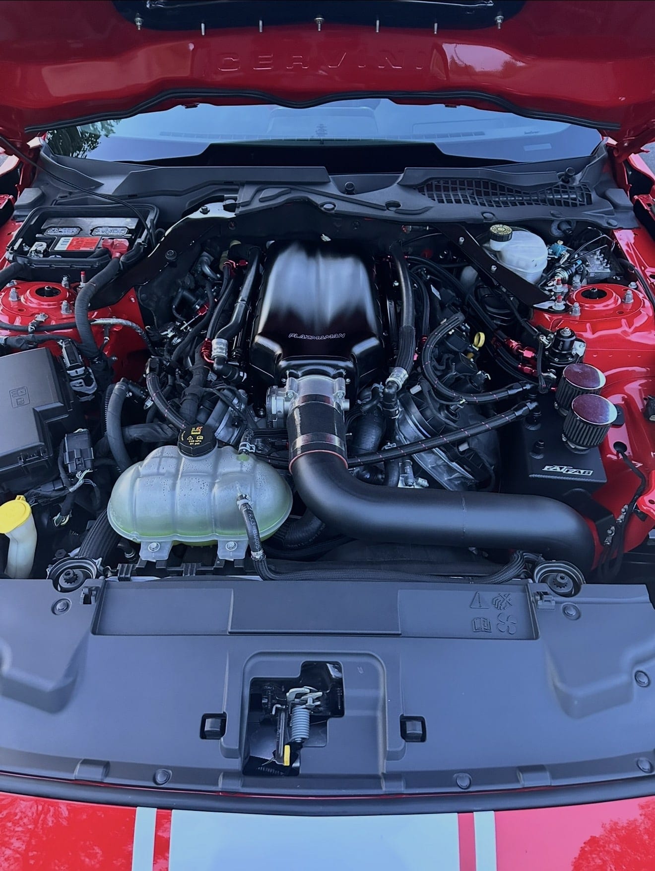 Engine bay of a red Mustang with an aftermarket intake manifold, black charge piping and red powder-coated strut towers