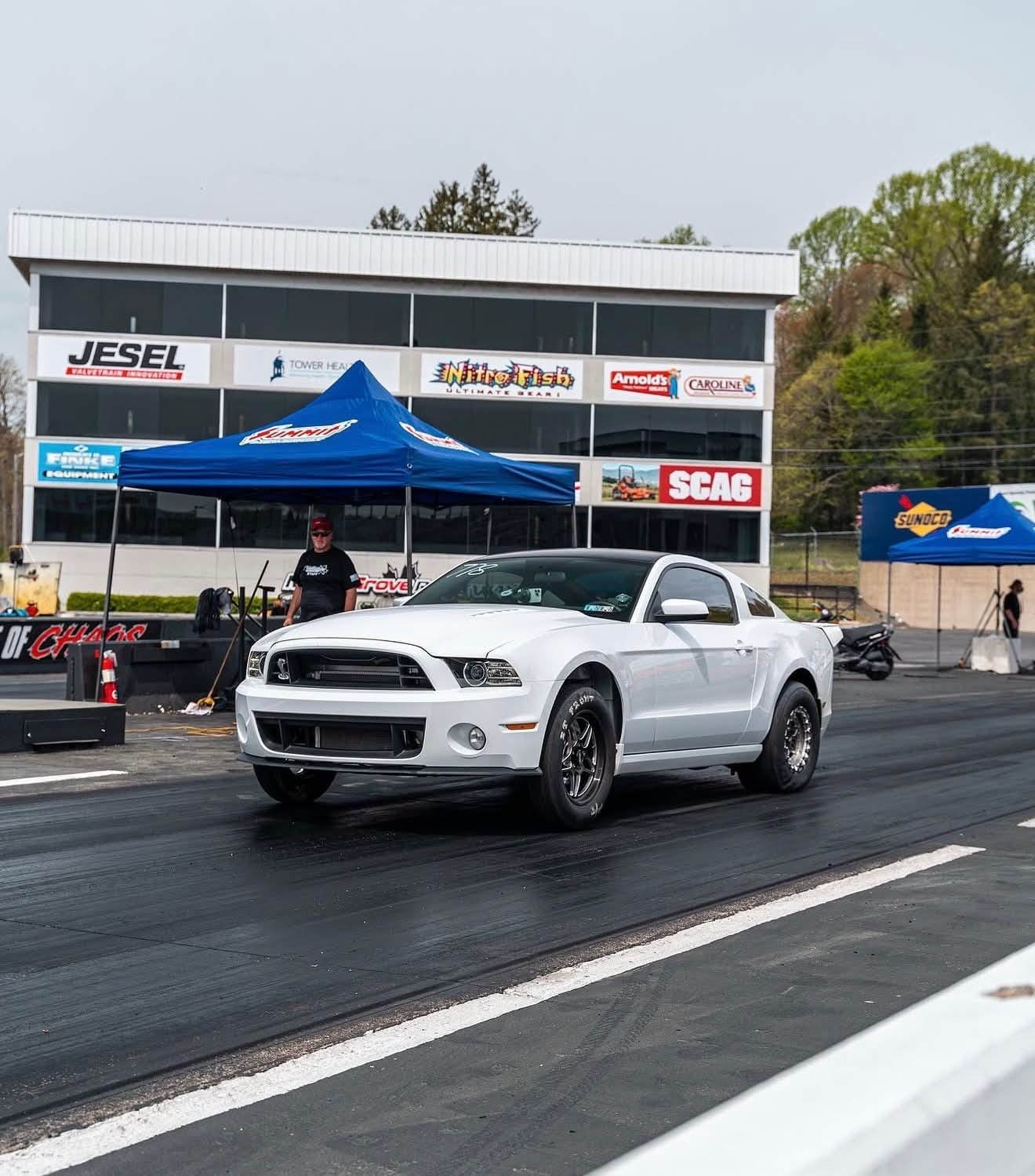A white Mustang with a front-mount intercooler and drag wheels staging at the start line of a drag strip