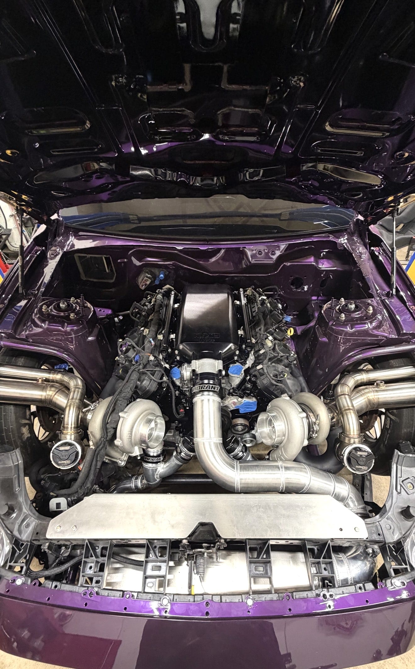 Engine bay of a deep purple build with a twin-turbocharged V8, stainless hot-side plumbing and polished charge piping